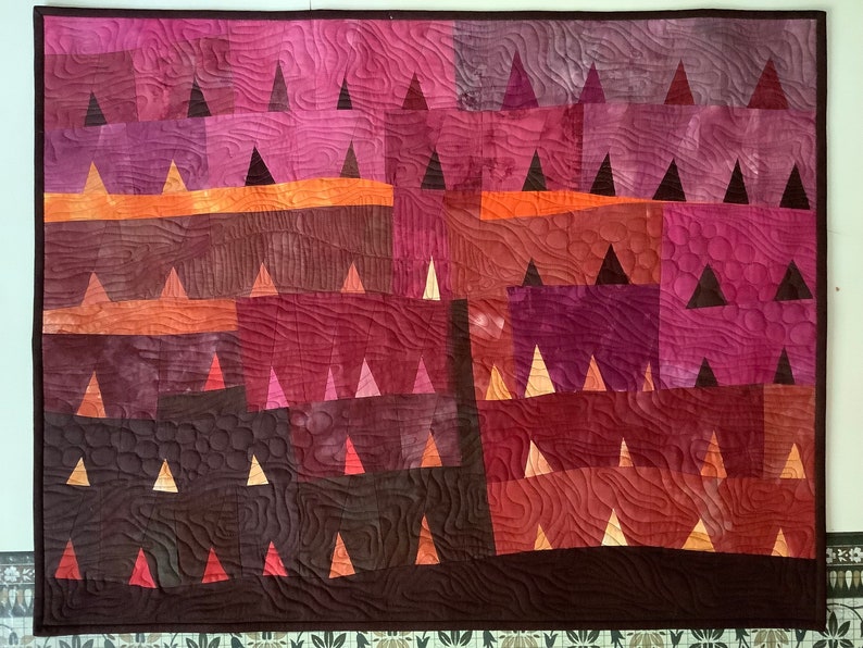 Op de afbeelding: Een quilt met een geometrisch patroon van driehoeken in tinten bruin, oranje en roze. De quilt is gemaakt van vierkante stoffen die aan elkaar zijn genaaid om een patchwork-ontwerp te cre&euml;ren.