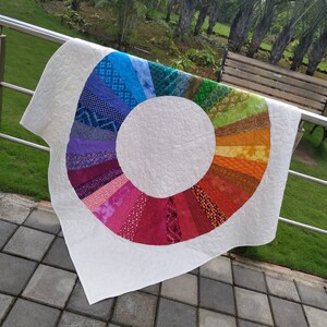 Op de afbeelding: Een wit quilt met een regenboogkleurige cirkel in het midden. De cirkel is gemaakt van verschillende stofresten die in een cirkelvormig patroon aan elkaar zijn genaaid. Het quilt hangt aan een leuning.