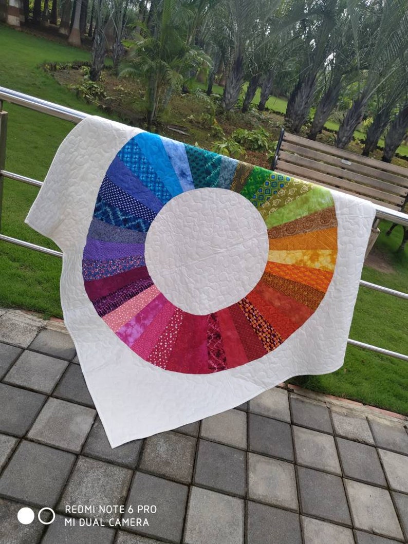 Op de afbeelding: Een wit quilt met een regenboogkleurige cirkel in het midden. De cirkel is gemaakt van verschillende stofresten die in een cirkelvormig patroon aan elkaar zijn genaaid. Het quilt hangt aan een leuning.