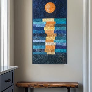 May include: A quilted wall hanging showing a night scene with a large orange moon and a path of light on the water. The artwork features a dark blue sky and water, with a textured, layered design. The piece is rectangular and mounted on a neutral wall.