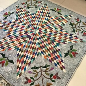 May include: A square quilt featuring a large central eight-pointed star composed of small, multi-colored diamond-shaped fabric pieces. The background is a light gray fabric with swirling quilted patterns and floral motifs in the corners.