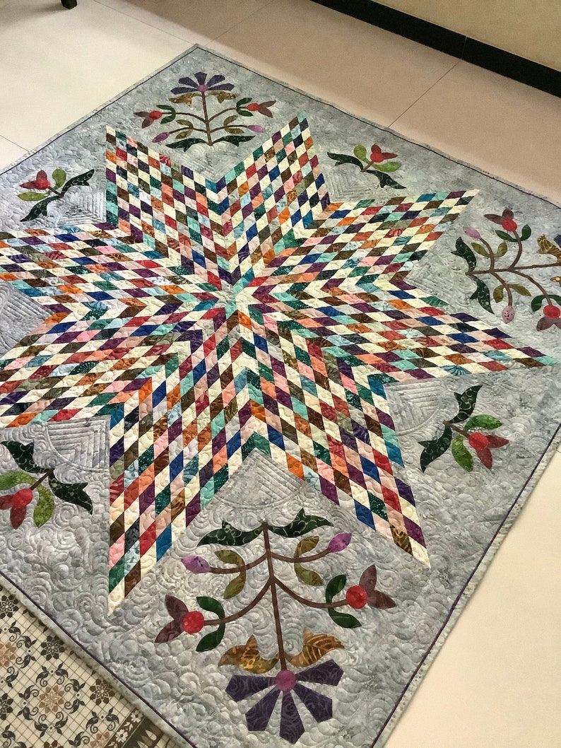 May include: A square quilt featuring a large central eight-pointed star composed of small, multi-colored diamond-shaped fabric pieces. The background is a light gray fabric with swirling quilted patterns and floral motifs in the corners.