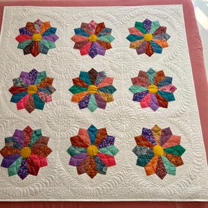 May include: A white quilt featuring nine colourful pinwheel blocks. Each block is made from a variety of fabrics in shades of pink, purple, blue, green, orange, and yellow. The quilt is quilted with a wavy pattern.