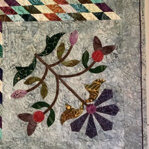 May include: A quilted fabric panel featuring a light gray background with a swirling stitched pattern. An appliqued branch design includes green leaves, pink buds, red berries, and a purple flower with petals made of various patterned fabrics.