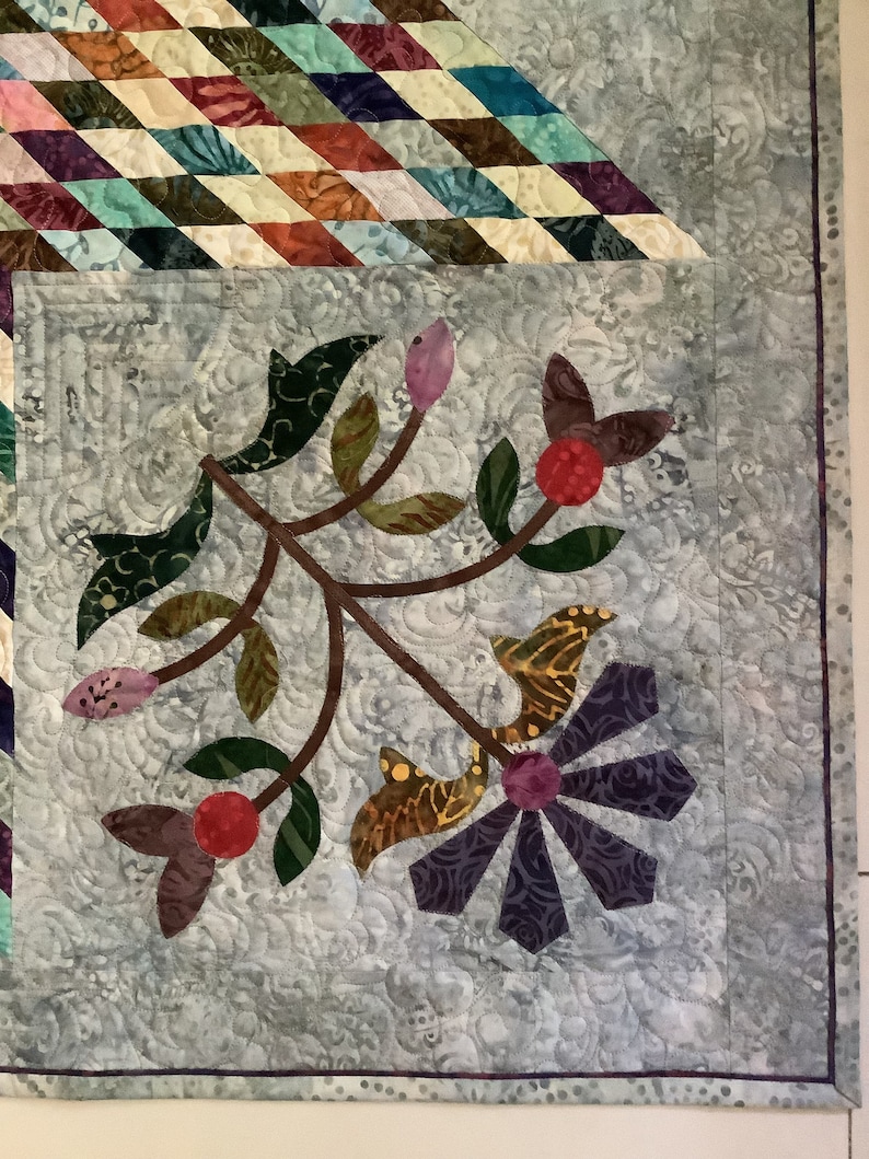 May include: A quilted fabric panel featuring a light gray background with a swirling stitched pattern. An appliqued branch design includes green leaves, pink buds, red berries, and a purple flower with petals made of various patterned fabrics.