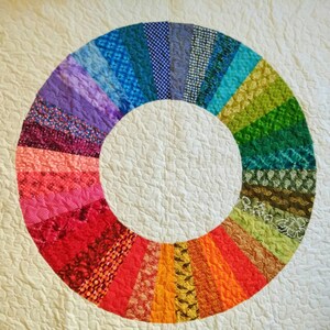 Op de afbeelding: Een wit quilt met een kleurrijk cirkelvormig ontwerp. De cirkel is gemaakt van verschillende stofresten in tinten paars, blauw, groen, rood, orange en geel. De stofresten zijn in een radiaal patroon gerangschikt, waardoor een regenboog-effect ontstaat.
