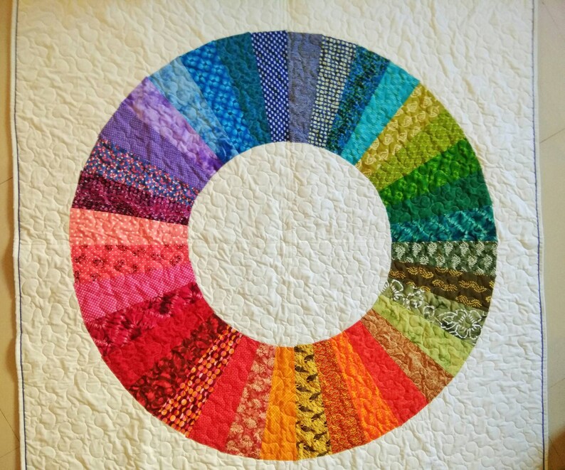 Op de afbeelding: Een wit quilt met een kleurrijk cirkelvormig ontwerp. De cirkel is gemaakt van verschillende stofresten in tinten paars, blauw, groen, rood, orange en geel. De stofresten zijn in een radiaal patroon gerangschikt, waardoor een regenboog-effect ontstaat.