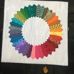Könnte beinhalten: Ein quadratischer, weißer Quilt mit einem kreisförmigen Design. Der Kreis besteht aus bunten, fächerförmigen Stoffstücken in einem Regenbogenmuster. Der Quilt hat eine gesteppte Textur.