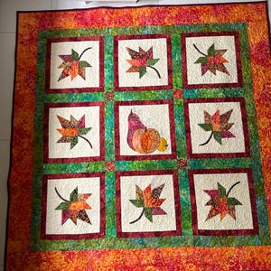 Op de afbeelding: Een vierkante quilt met een raster van negen blokken en een donkerrode en groene rand. Acht blokken tonen kleurrijke patchwork herfstbladeren op een crèmekleurige achtergrond, terwijl het middelste blok een peer, een pompoen en een kalebas toont.