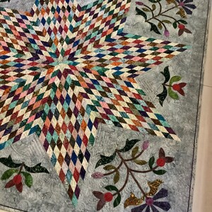 May include: A quilt featuring a large central star composed of multicolored diamond-shaped fabric patches. The background is a light gray fabric with a swirling texture, decorated with floral appliqu&eacute;s in shades of green, red, and purple.