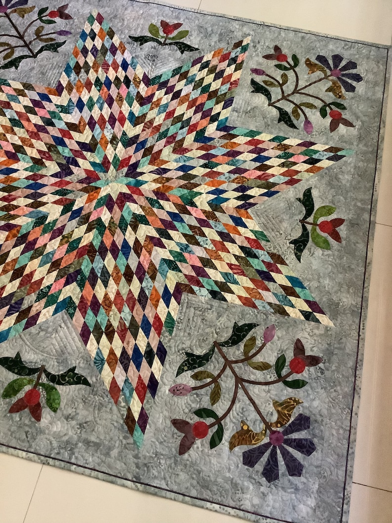 May include: A quilt featuring a large central star composed of multicolored diamond-shaped fabric patches. The background is a light gray fabric with a swirling texture, decorated with floral appliqu&eacute;s in shades of green, red, and purple.