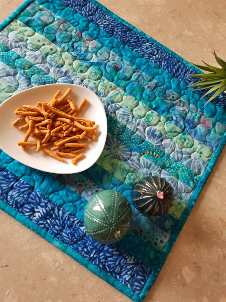 May include: A blue and green quilted table runner with a white plate of brown and orange snacks, a green ceramic ornament, and a blue ceramic ornament.