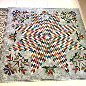 May include: A square quilt featuring a large central eight-pointed star composed of multicolored diamond-shaped fabric pieces. The background is a light gray fabric with swirling quilted patterns, accented by floral motifs in the corners.