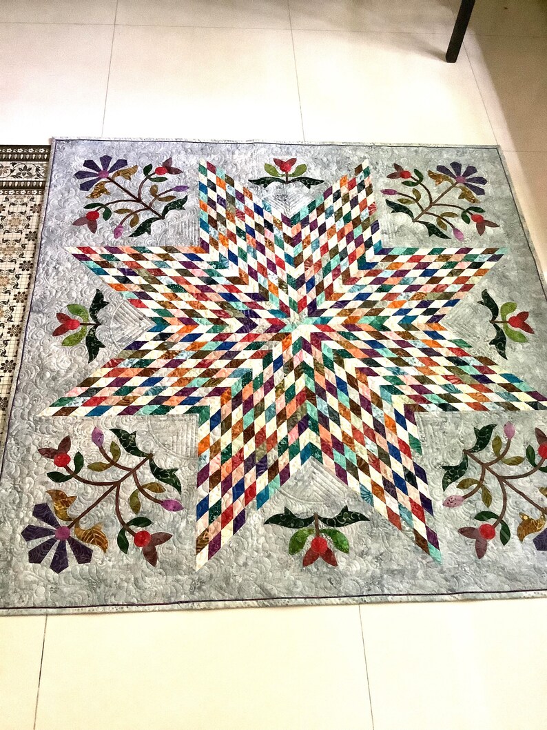 May include: A square quilt featuring a large central eight-pointed star composed of multicolored diamond-shaped fabric pieces. The background is a light gray fabric with swirling quilted patterns, accented by floral motifs in the corners.