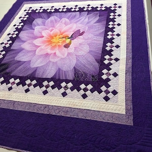 May include: A purple and white quilt with a large flower and hummingbird design in the center. The quilt is quilted with a floral pattern and has a purple border.