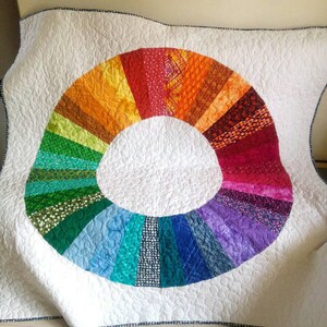 Op de afbeelding: Een wit quilt met een kleurrijk regenboogcirkelontwerp. De cirkel is gemaakt van verschillende stofresten in tinten rood, oranje, geel, groen, blauw, indigo en violet.