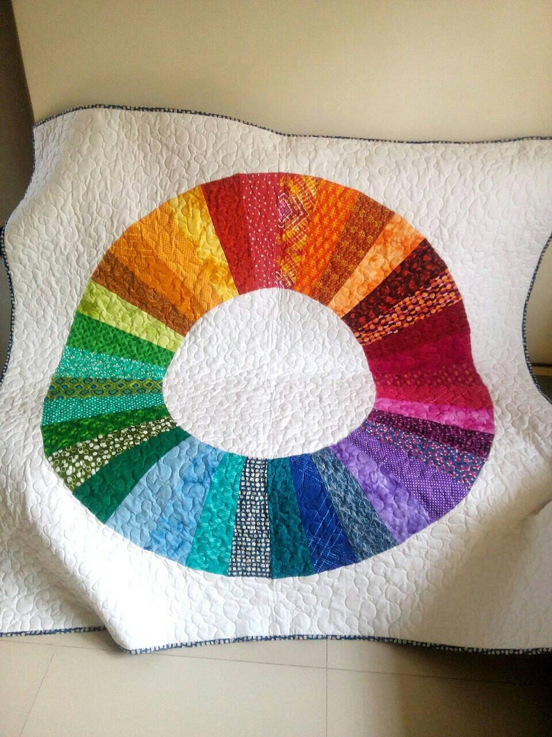 Op de afbeelding: Een wit quilt met een kleurrijk regenboogcirkelontwerp. De cirkel is gemaakt van verschillende stofresten in tinten rood, oranje, geel, groen, blauw, indigo en violet.