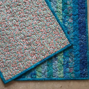 May include: Two quilted placemats with a blue border. One placemat has a white background with a small red and blue floral pattern. The other placemat has a blue and green striped pattern.