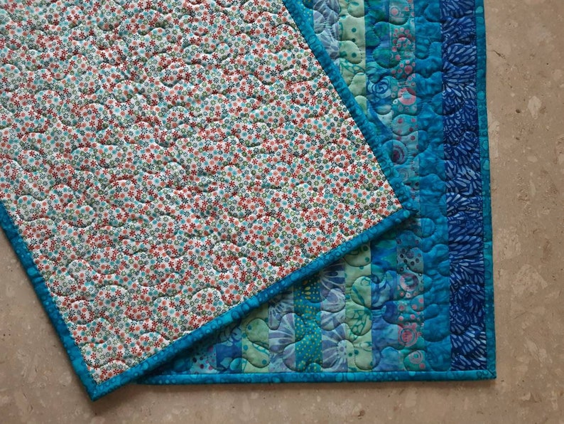 May include: Two quilted placemats with a blue border. One placemat has a white background with a small red and blue floral pattern. The other placemat has a blue and green striped pattern.