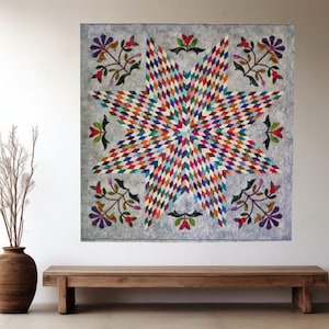 May include: A square wall quilt featuring a large central eight-pointed star composed of small, multi-colored diamond-shaped fabric pieces. The background is a light gray fabric, accented with floral motifs in the corners and along the edges.