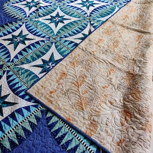 May include: A blue and white quilt with a star pattern on the front and a cream-colored backing with a floral pattern. The quilt is folded in half, revealing the backing.