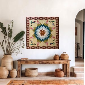 May include: A colorful quilt with a starburst design hangs on a white wall. The quilt features a cream-colored background with a central star in shades of blue, green, and brown. Decorative baskets and a wooden bench are in front of the wall.