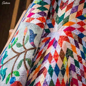 May include: A quilt featuring a vibrant diamond pattern in various colors including red, orange, green, blue, and purple. One section displays a light gray fabric with an embroidered branch, leaves, and flower motif. The word Sakura is visible.
