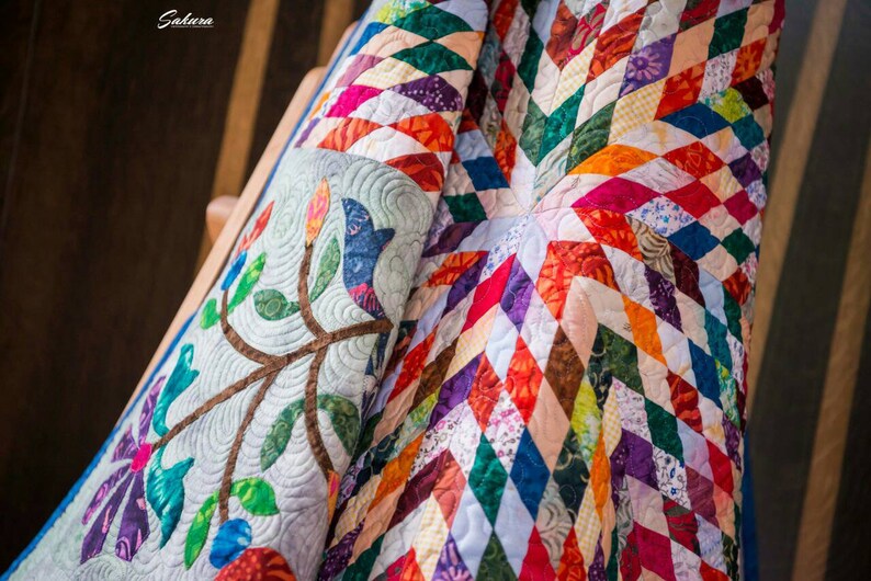 May include: A quilt featuring a vibrant diamond pattern in various colors including red, orange, green, blue, and purple. One section displays a light gray fabric with an embroidered branch, leaves, and flower motif. The word Sakura is visible.