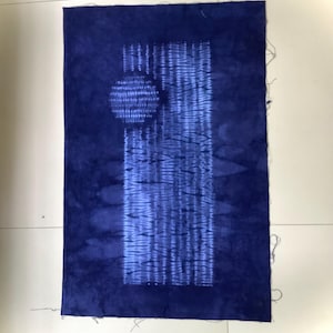 May include: A rectangular piece of fabric dyed in a deep indigo blue. The centre features a vertical panel with a lighter blue shibori-style resist pattern, including a circular motif at the top left and horizontal dashed lines throughout the panel.