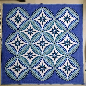 May include: A blue and white quilt with a geometric pattern of stars and circles. The quilt is made of fabric squares sewn together to create a larger design.