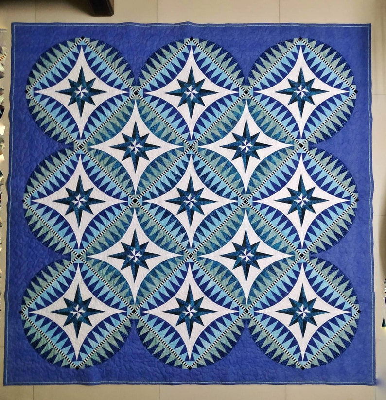 May include: A blue and white quilt with a geometric pattern of stars and circles. The quilt is made of fabric squares sewn together to create a larger design.