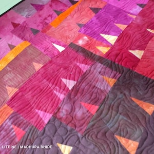 Op de afbeelding: Een quilt met een geometrisch patroon van driehoeken in tinten roze, oranje en paars. De quilt is gestikt met een golvend patroon.