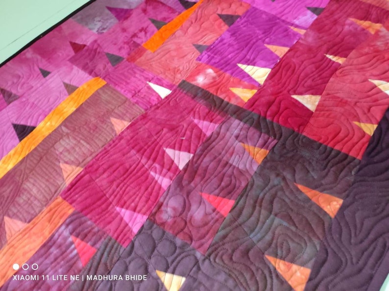 Op de afbeelding: Een quilt met een geometrisch patroon van driehoeken in tinten roze, oranje en paars. De quilt is gestikt met een golvend patroon.