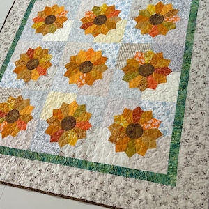 May include: Handmade quilt featuring nine sunflower appliqués. Each sunflower has a brown center and petals in shades of orange, yellow, and red. The quilt has a white background with floral and geometric patterns, and a green border.