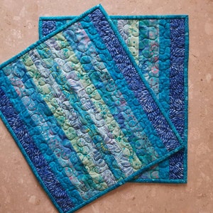 May include: Two blue and green quilted placemats with a striped pattern. The placemats are made of fabric scraps and have a unique, handmade look.