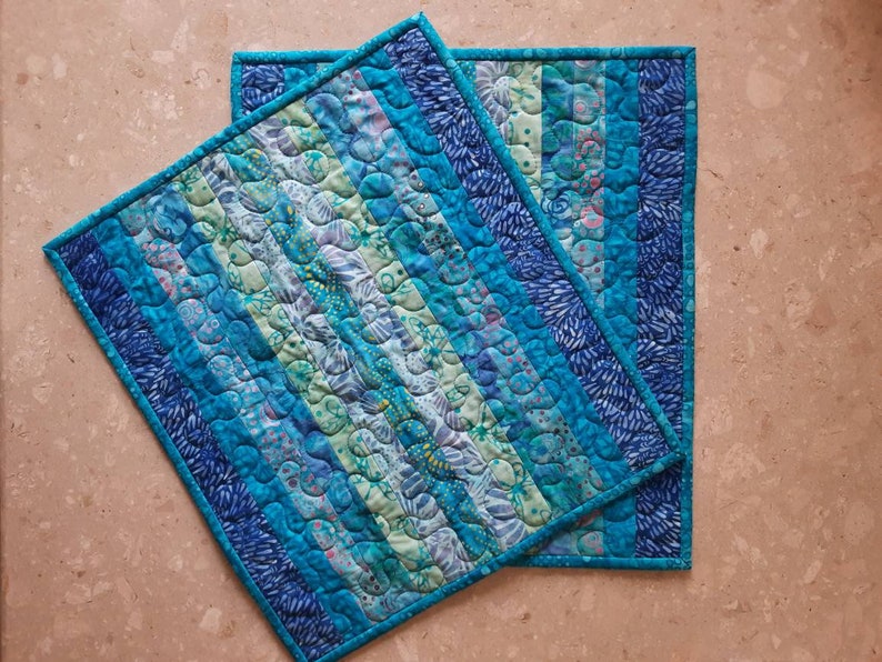 May include: Two blue and green quilted placemats with a striped pattern. The placemats are made of fabric scraps and have a unique, handmade look.