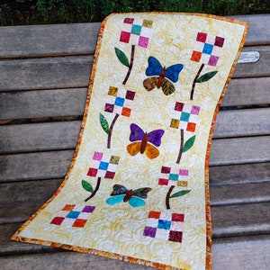 May include: A yellow table runner with a quilted design featuring colorful butterflies and flowers. The runner has a brown stem and green leaves. The runner has a brown and orange border.