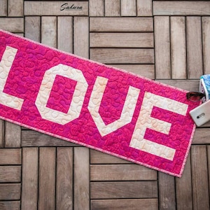May include: A pink quilted fabric banner with the word "LOVE" in white letters. The banner is lying on a wooden surface.