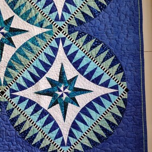 May include: A blue and white quilt with a starburst pattern in the center. The quilt is made of various shades of blue and green fabric and has a black and white border.