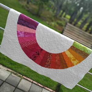 Op de afbeelding: Een wit quilt met een kleurrijk regenboogvormig ontwerp. De boog is gemaakt van verschillende stofresten in tinten roze, rood, oranje, geel, groen en paars. Het quilt hangt aan een metalen leuning.