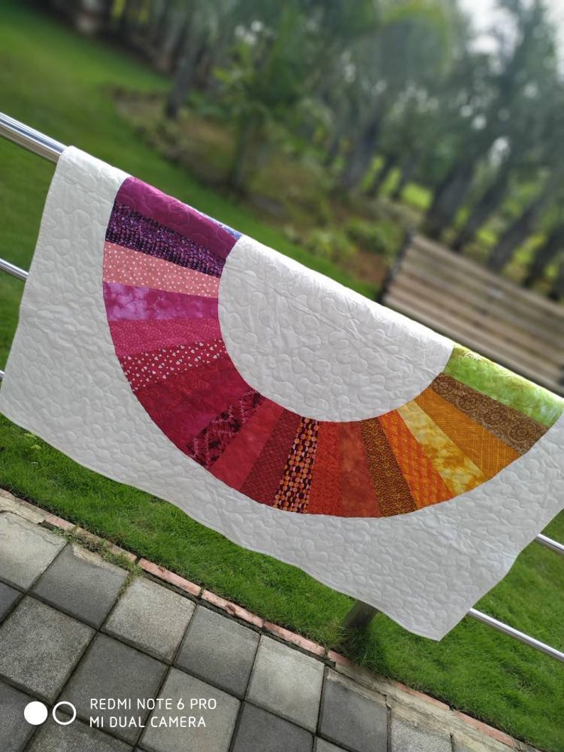 Op de afbeelding: Een wit quilt met een kleurrijk regenboogvormig ontwerp. De boog is gemaakt van verschillende stofresten in tinten roze, rood, oranje, geel, groen en paars. Het quilt hangt aan een metalen leuning.