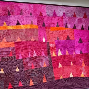 Op de afbeelding: Een quilt met een geometrisch patroon van driehoeken in tinten roze, rood, oranje en paars. De quilt is gestikt met een golvend patroon.