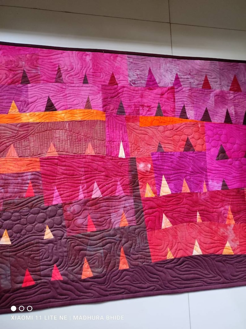 Op de afbeelding: Een quilt met een geometrisch patroon van driehoeken in tinten roze, rood, oranje en paars. De quilt is gestikt met een golvend patroon.