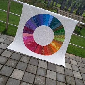 Op de afbeelding: Een wit quilt met een kleurrijk cirkelontwerp in het midden. De cirkel is gemaakt van verschillende gekleurde stofstroken die in een regenboogpatroon zijn gerangschikt.
