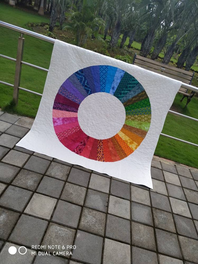 Op de afbeelding: Een wit quilt met een kleurrijk cirkelontwerp in het midden. De cirkel is gemaakt van verschillende gekleurde stofstroken die in een regenboogpatroon zijn gerangschikt.