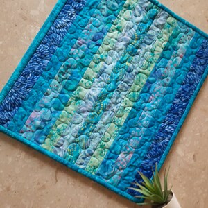 May include: A blue and green quilted throw pillow with a wavy pattern. The pillow is made of fabric with a variety of blue and green shades.