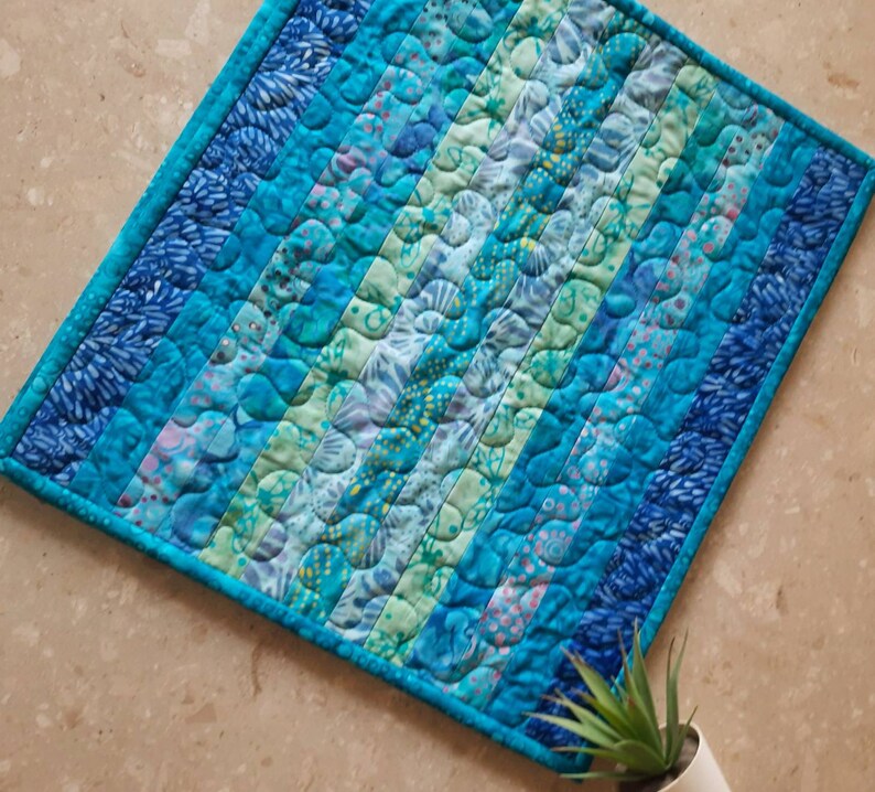May include: A blue and green quilted throw pillow with a wavy pattern. The pillow is made of fabric with a variety of blue and green shades.