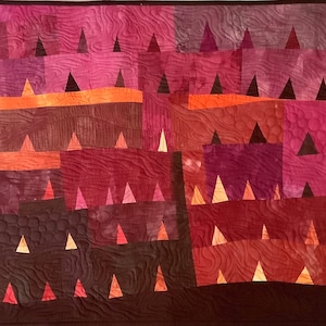 Op de afbeelding: Een quilt met een geometrisch patroon van driehoeken in tinten bruin, rood en oranje. De driehoeken zijn gerangschikt in rijen en kolommen, waardoor een gevoel van diepte en beweging ontstaat.