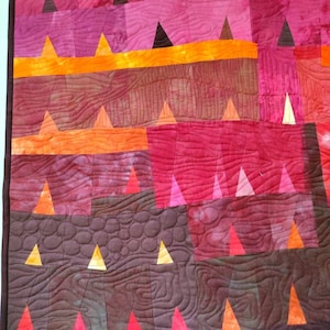 Op de afbeelding: Een quilt met een patchwork-ontwerp met verschillende tinten rood, oranje en bruin. De quilt is gemaakt van driehoeken en vierkanten, en de stiksels zijn zichtbaar. De quilt is gestikt met een patroon van cirkels en lijnen.