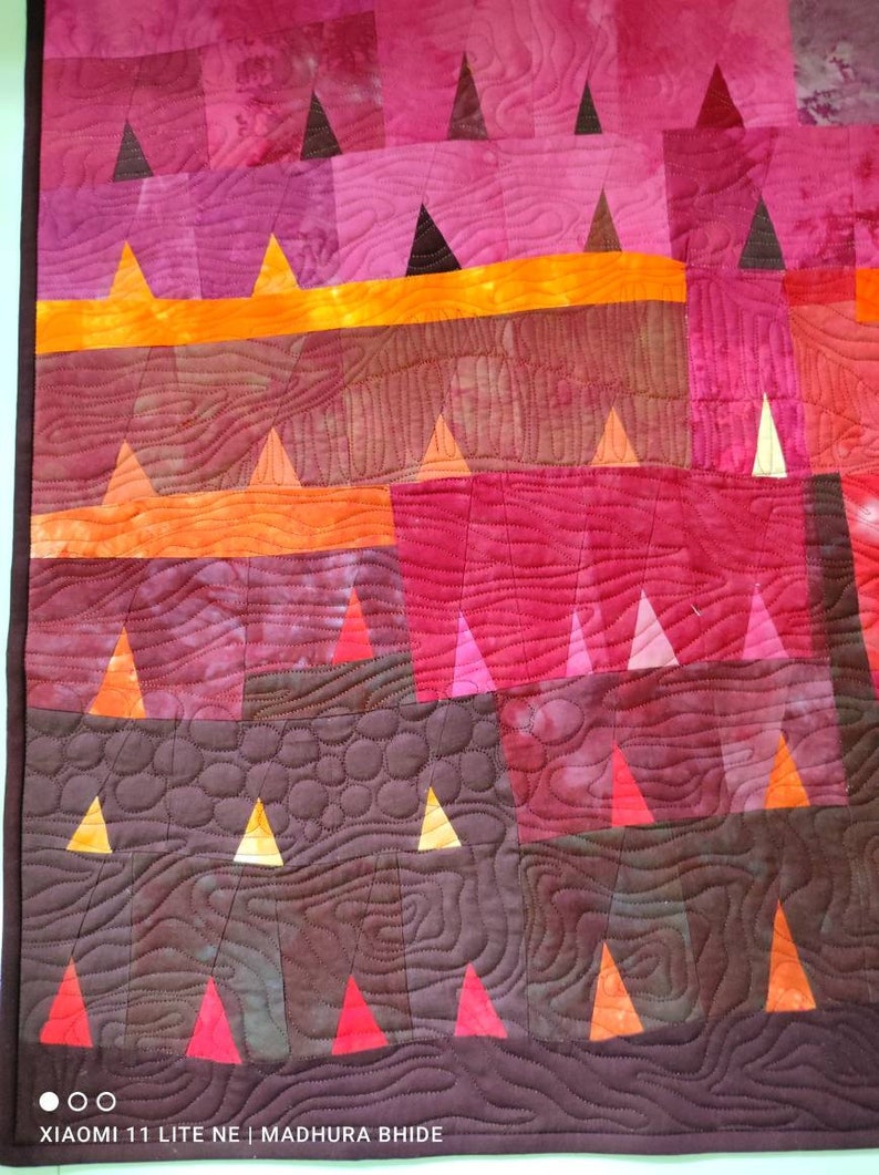 Op de afbeelding: Een quilt met een patchwork-ontwerp met verschillende tinten rood, oranje en bruin. De quilt is gemaakt van driehoeken en vierkanten, en de stiksels zijn zichtbaar. De quilt is gestikt met een patroon van cirkels en lijnen.